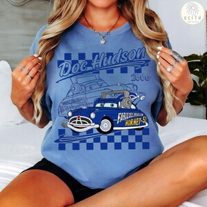 May include: A light blue t-shirt featuring a cartoon image of a blue car with the text "Doc Hudson 2006" and "Fabulous Hudson Hornet 51". The design includes a checkered pattern and a car blueprint.