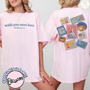 May include: Light pink t-shirt with the text "wish you were here" and "Walt Disney World" in blue. The back of the shirt features various Disney-themed postage stamp designs. The shirt is labeled "Comfort Colors Blossom".