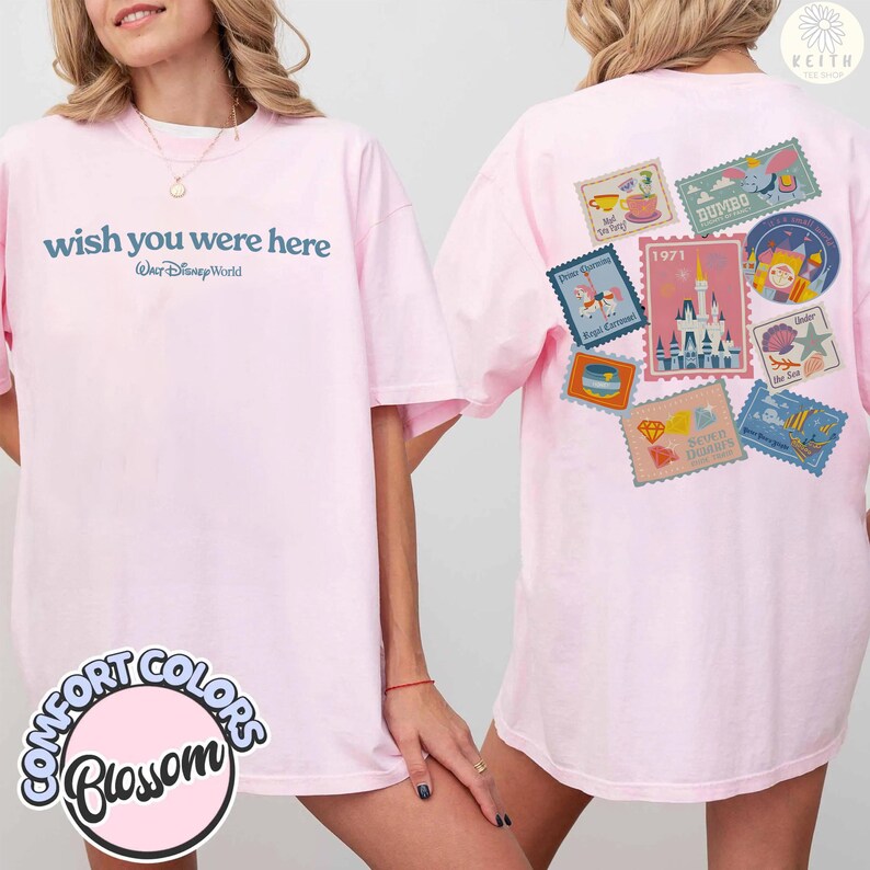 May include: Light pink t-shirt with the text "wish you were here" and "Walt Disney World" in blue. The back of the shirt features various Disney-themed postage stamp designs. The shirt is labeled "Comfort Colors Blossom".