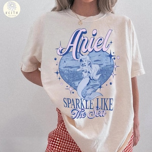 Puede incluir: Camiseta color crema con un gr&aacute;fico de Ariel la sirenita dentro de un coraz&oacute;n azul. El texto "Ariel" est&aacute; encima del coraz&oacute;n y "Sparkle Like The Sea" debajo. La camiseta tiene mangas cortas.
