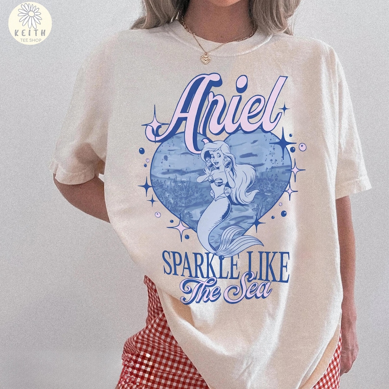 Puede incluir: Camiseta color crema con un gr&aacute;fico de Ariel la sirenita dentro de un coraz&oacute;n azul. El texto "Ariel" est&aacute; encima del coraz&oacute;n y "Sparkle Like The Sea" debajo. La camiseta tiene mangas cortas.
