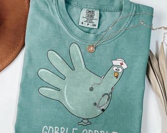 May include: A light green short-sleeve t-shirt featuring a graphic of a turkey shaped like a hand, wearing a nurse cap and a stethoscope. The text GOBBLE GOBBLE is printed below the graphic. The shirt has a Comfort Colors brand label.