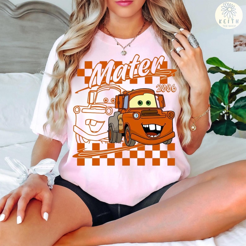 May include: A light pink t-shirt featuring a cartoon image of Mater from the movie Cars. The image includes the word "Mater" in orange lettering, the year "2006", and a checkered background.