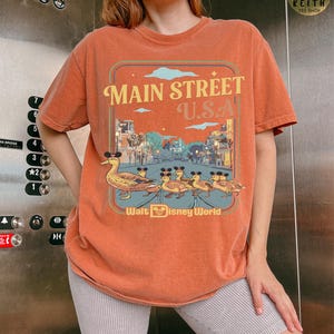 May include: Orange t-shirt with a graphic of ducks wearing mouse ears walking down Main Street U.S.A. The text "Main Street U.S.A." and "Walt Disney World" are also displayed on the shirt.