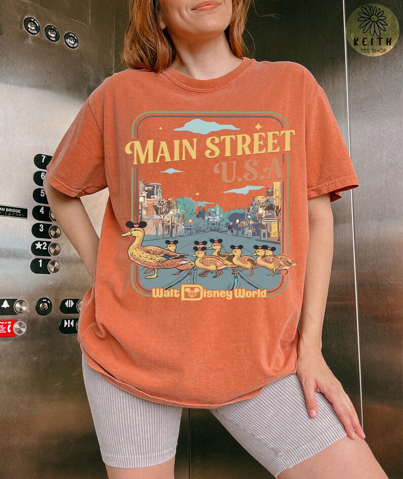 May include: Orange t-shirt with a graphic of ducks wearing mouse ears walking down Main Street U.S.A. The text "Main Street U.S.A." and "Walt Disney World" are also displayed on the shirt.