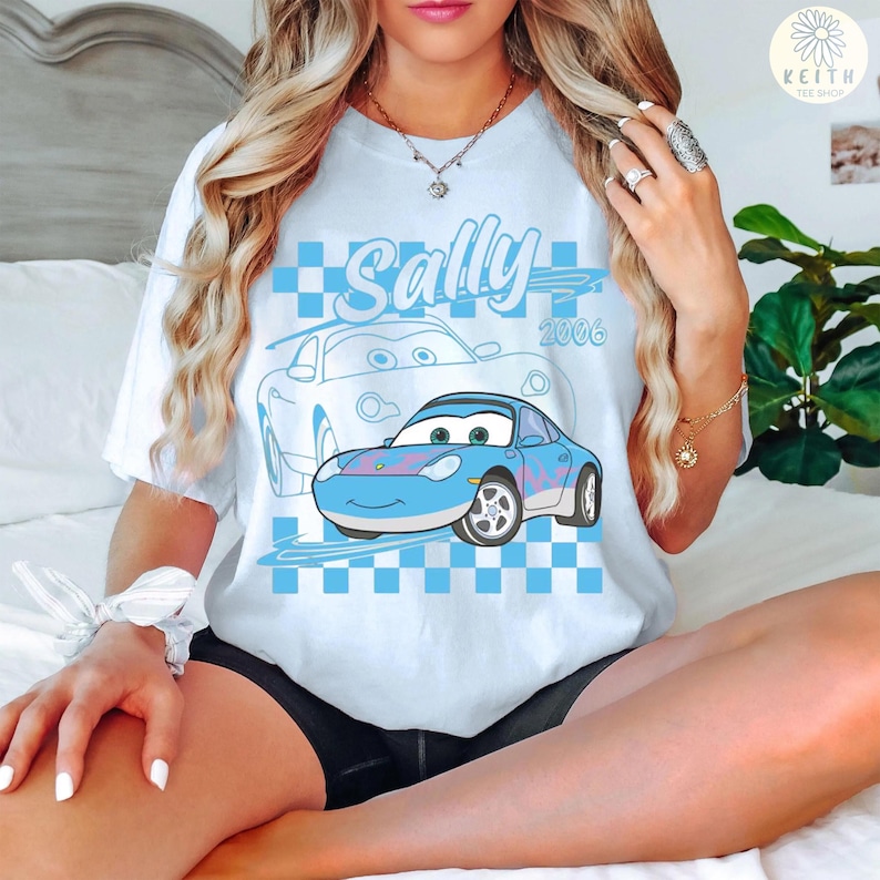 May include: Light blue t-shirt featuring a cartoon image of Sally Carrera from the movie Cars. The design includes the name "Sally" in a script font, the year "2006", and a checkered pattern.