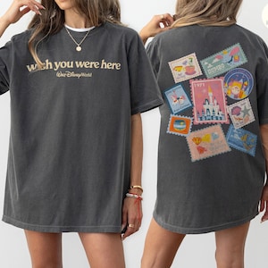 May include: Dark gray t-shirt with the text "wish you were here" and "Walt Disney World" in gold lettering. The back of the shirt features colorful postage stamp designs with various Disney-themed illustrations.