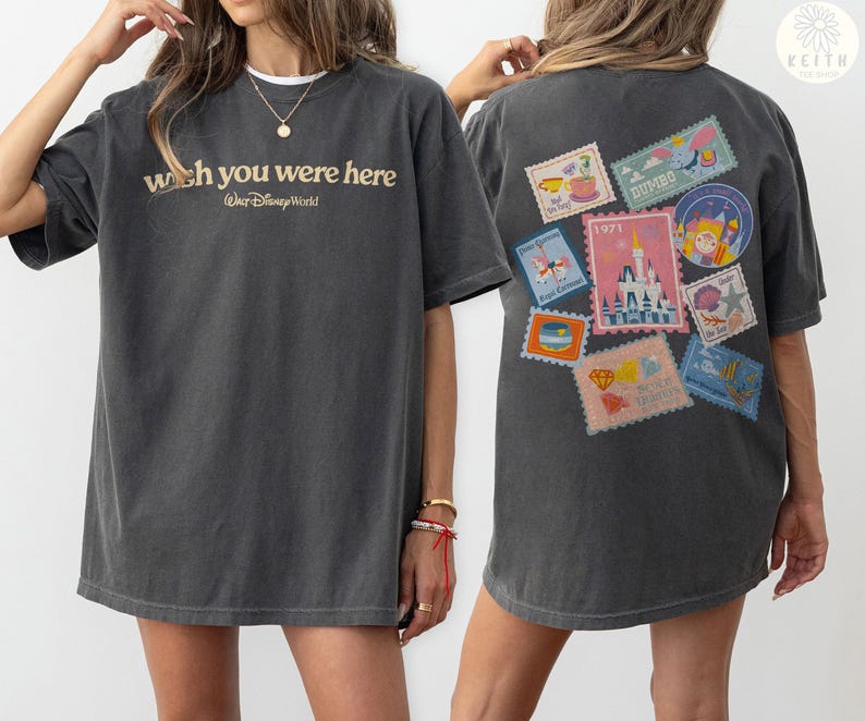 May include: Dark gray t-shirt with the text "wish you were here" and "Walt Disney World" in gold lettering. The back of the shirt features colorful postage stamp designs with various Disney-themed illustrations.