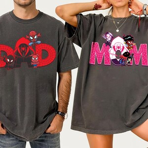 May include: Two dark gray t-shirts. One features the word "DAD" in red with Spider-Man characters. The other shirt has the word "MOM" in pink with Spider-Verse characters. Both shirts are oversized and have short sleeves.