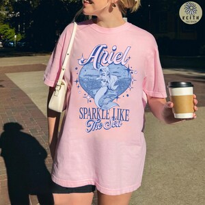 Puede incluir: Camiseta rosa claro con un gr&aacute;fico de Ariel la sirena dentro de un coraz&oacute;n azul y el texto "Sparkle Like The Sea". La camiseta es de Keith Tee Shop.