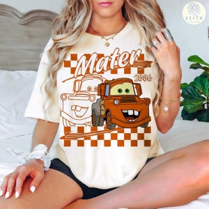 May include: Cream-colored t-shirt featuring a cartoon image of Mater from the movie Cars. The design includes the name "Mater" in a stylized font, the year "2006", and a checkered pattern in shades of brown and orange.