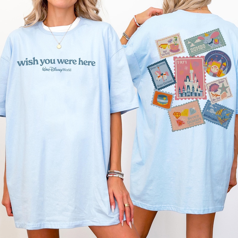 May include: Light blue t-shirt with the text "wish you were here" and "Walt Disney World" printed on the front. The back of the shirt features various colorful postage stamp designs with Disney themes.