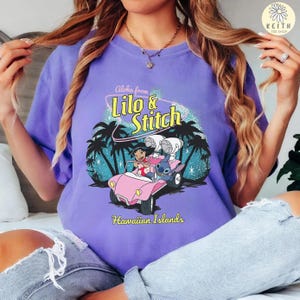 May include: A purple t-shirt featuring a graphic of Lilo and Stitch in a pink car, with the text "Aloha from Lilo & Stitch" and "Hawaiian Islands". The design includes palm trees and a tropical scene.