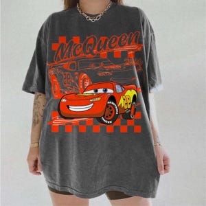 May include: Dark gray t-shirt featuring a graphic of Lightning McQueen from the movie Cars. The design includes the text "McQueen 2006" and a checkered flag pattern. The shirt is oversized and has short sleeves.