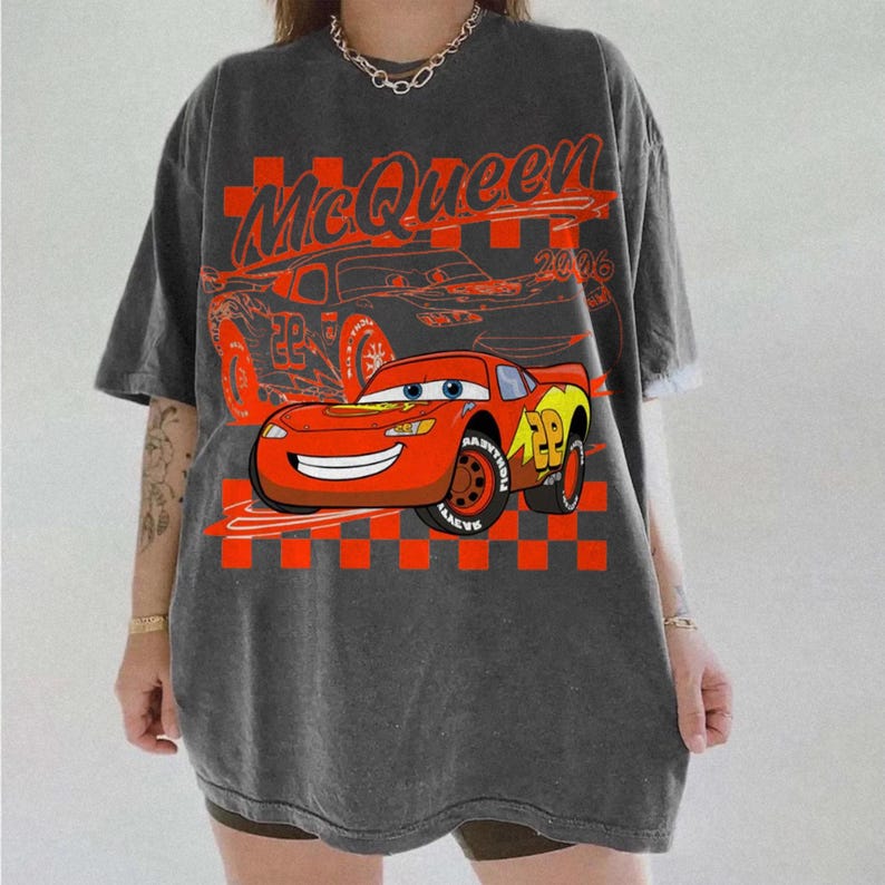 May include: Dark gray t-shirt featuring a graphic of Lightning McQueen from the movie Cars. The design includes the text "McQueen 2006" and a checkered flag pattern. The shirt is oversized and has short sleeves.