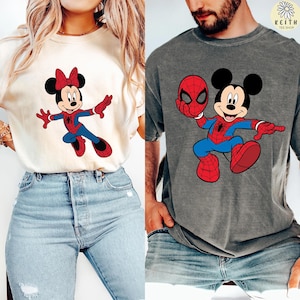 Może przedstawiać: Dwie koszulki z postaciami z kreskówek. Jedna to kremowa koszulka z Minnie Mouse w kostiumie Spider-Mana. Druga to szara koszulka z Myszka Miki w kostiumie Spider-Mana trzymającego maskę Spider-Mana. Koszulki pochodzą ze sklepu Keith Tee Shop.