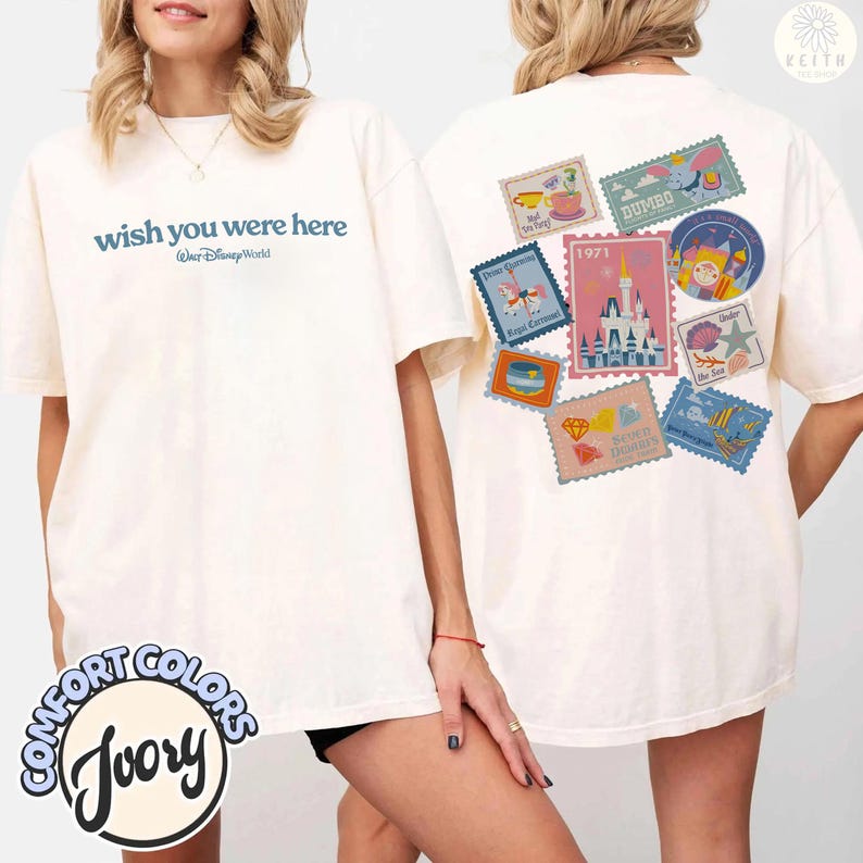May include: Ivory-colored t-shirt with the text "wish you were here" and "Walt Disney World" in blue. The back features various colorful postage stamp designs with Disney themes. The shirt is from Comfort Colors.
