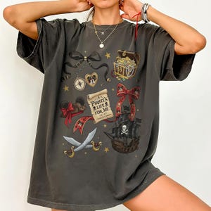 Könnte beinhalten: Dunkelgraues T-Shirt mit Piratenmotiven. Das Design umfasst eine Schatzkiste, ein Piratenschiff, Schwerter, Mickey Mouse Ohren und den Text "Pirate's Life For Me". Das Shirt hat eine lockere Passform.