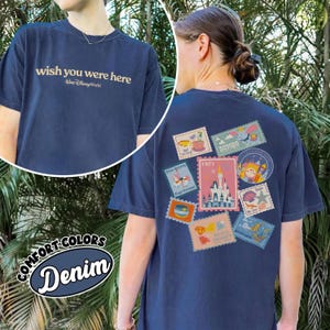 May include: Navy blue t-shirt with "wish you were here" text on the front and a collection of colorful postage stamp designs on the back. The stamps feature various Disney-themed images, including a castle and Dumbo. The shirt is labeled "Comfort Colors Denim".