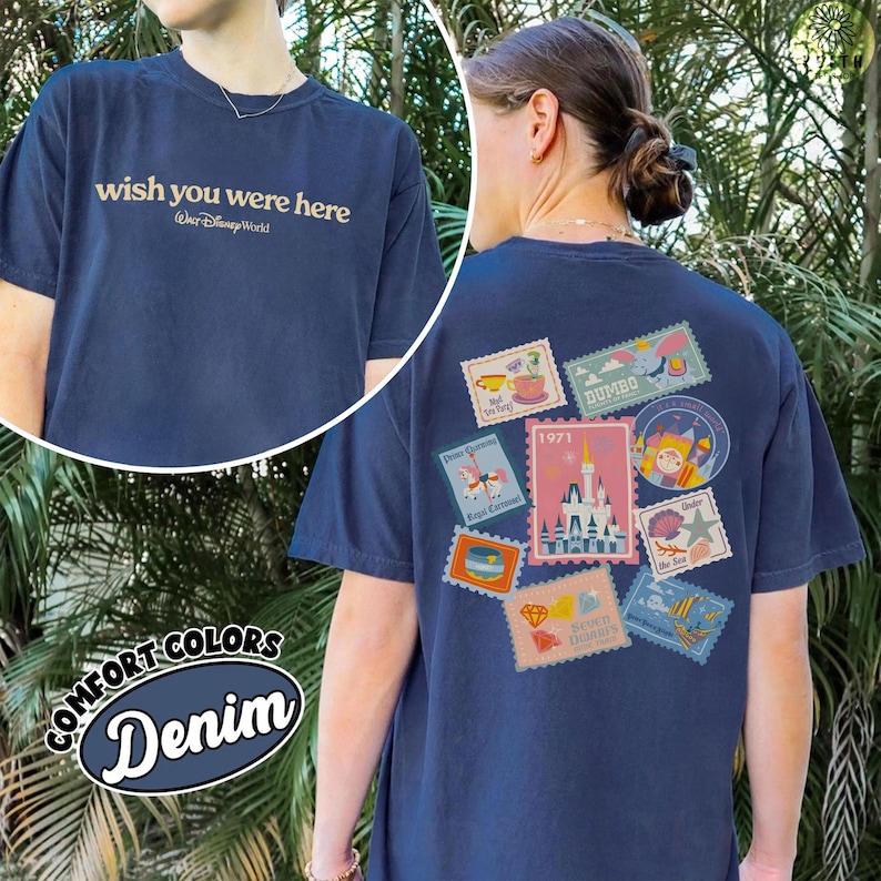 May include: Navy blue t-shirt with "wish you were here" text on the front and a collection of colorful postage stamp designs on the back. The stamps feature various Disney-themed images, including a castle and Dumbo. The shirt is labeled "Comfort Colors Denim".