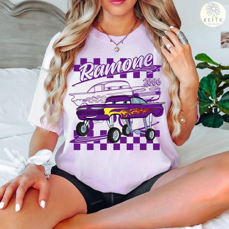 May include: A light purple t-shirt featuring a graphic of a purple car with flames and the name "Ramone 2006" in white. The design is set against a purple and white checkered background. The shirt has short sleeves.