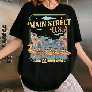 May include: Black t-shirt with a vintage-style design of Main Street U.S.A. featuring a duck family wearing hats. The graphic includes the text "Main Street U.S.A." and "Walt Disney World".