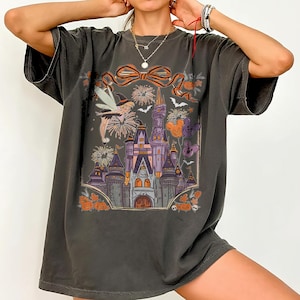 May include: Dark gray t-shirt featuring a colorful graphic of a castle, fireworks, and Halloween-themed elements. The design includes a fairy, pumpkins, and Mickey Mouse-shaped balloons. The shirt has a relaxed fit and short sleeves.
