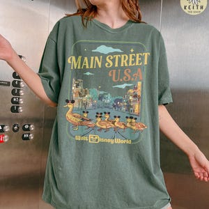 May include: Green t-shirt with a graphic showing "Main Street U.S.A." above a street scene and ducks wearing Mickey Mouse ears. The text "Walt Disney World" is below the graphic.