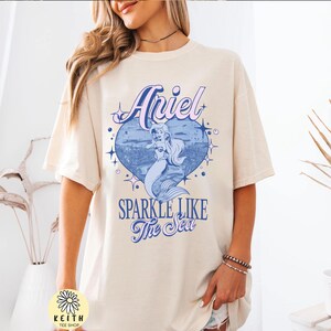 Puede incluir: Camiseta color crema con un gr&aacute;fico de Ariel la sirenita dentro de un coraz&oacute;n azul con el texto "Ariel" y "Sparkle Like The Sea". La camiseta tiene mangas cortas y un corte holgado.