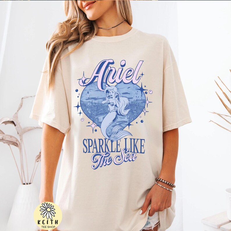 Puede incluir: Camiseta color crema con un gr&aacute;fico de Ariel la sirenita dentro de un coraz&oacute;n azul con el texto "Ariel" y "Sparkle Like The Sea". La camiseta tiene mangas cortas y un corte holgado.