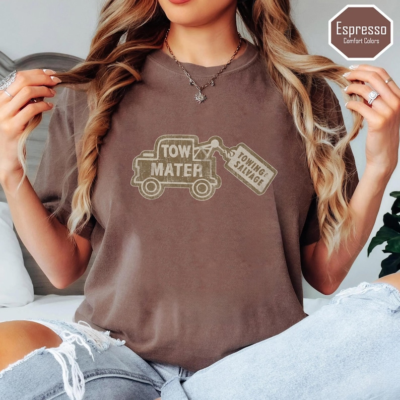 May include: Espresso-colored t-shirt featuring a distressed graphic of a tow truck with the words "Tow Mater" and "Towing & Salvage." The shirt is a comfort color and has a relaxed fit. The shirt is a brown color.