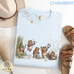 May include: Light blue t-shirt with a cartoon illustration of Ewoks in a forest setting. The Ewoks are depicted with spears, slingshots, and a Stormtrooper helmet. The shirt has the word "CHAMBRAY" printed at the top.