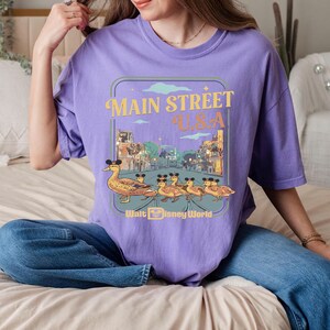May include: A lilac t-shirt with a graphic of Main Street U.S.A. and ducks wearing Mickey Mouse ears. The design has a retro style and the text "Walt Disney World".
