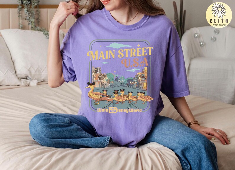 May include: A lilac t-shirt with a graphic of Main Street U.S.A. and ducks wearing Mickey Mouse ears. The design has a retro style and the text "Walt Disney World".