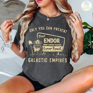Puede incluir: Camiseta gris oscuro con el texto "ONLY YOU CAN PREVENT" sobre un letrero que dice "Visit ENDOR Hiking-Camping-Feasting Lunar Forest GALACTIC EMPIRES". El letrero presenta un personaje de dibujos animados con una pala.