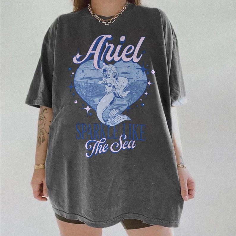 Puede incluir: Camiseta gris oscuro de gran tama&ntilde;o con el nombre "Ariel" en escritura cursiva sobre un gr&aacute;fico azul en forma de coraz&oacute;n. El gr&aacute;fico representa una sirena y el texto "Sparkle Like The Sea".