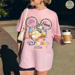 May include: A light pink t-shirt featuring a Disney design with the text "Disney 2026". The design includes the characters from Beauty and the Beast, fireworks, and Mickey Mouse ears. The word "Name" is also included in the design.