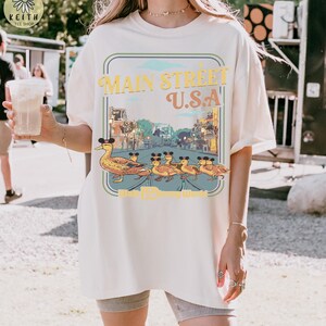 May include: Off-white t-shirt with a graphic of ducks wearing Mickey Mouse ears, walking in a line. The graphic reads "Main Street U.S.A." in yellow and orange, with a blue and green border. The shirt has a loose fit.
