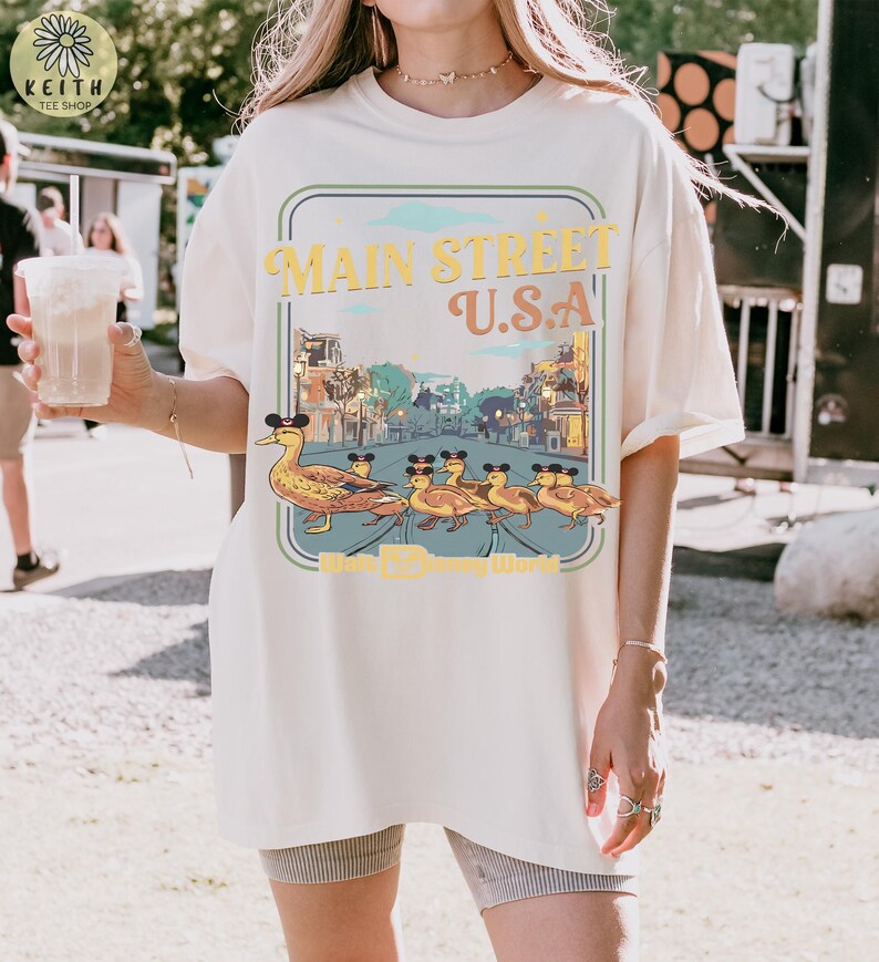 May include: Off-white t-shirt with a graphic of ducks wearing Mickey Mouse ears, walking in a line. The graphic reads "Main Street U.S.A." in yellow and orange, with a blue and green border. The shirt has a loose fit.