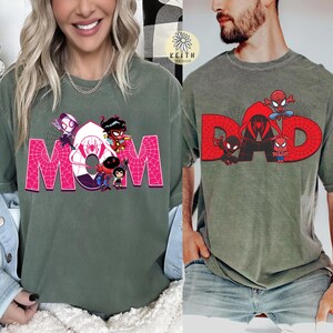 May include: Two olive green t-shirts. One features the word "MOM" in pink, with a spiderweb design and cartoon characters. The other shirt says "DAD" in red, with a spiderweb design and cartoon characters.