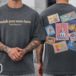 May include: Dark gray t-shirt with "wish you were here" and "Walt Disney World" text. The back features colorful postage stamp designs with Disney themes. The shirt is a Pepper Comfort Colors brand.