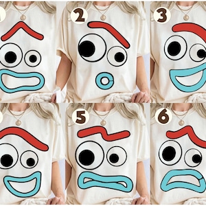 May include: A collection of six cream-colored t-shirts, each featuring a different cartoon face design. The faces have black eyes, red eyebrows, and blue mouths. The designs showcase a range of emotions, from happy to sad.