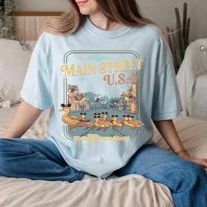 May include: Pale blue t-shirt with a graphic of ducks wearing Mickey Mouse ears, walking in front of a Main Street U.S.A. scene. The text "Main Street U.S.A. Walt Disney World" is also printed on the shirt.