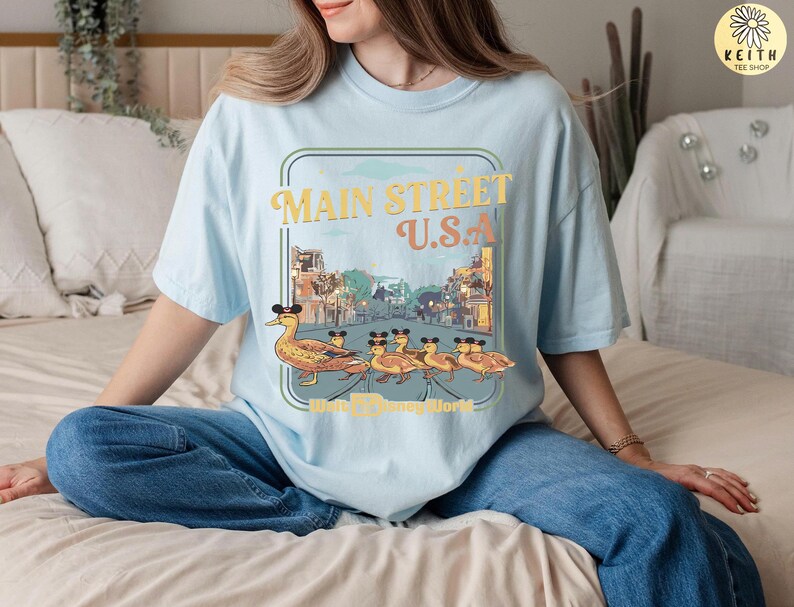 May include: Pale blue t-shirt with a graphic of ducks wearing Mickey Mouse ears, walking in front of a Main Street U.S.A. scene. The text "Main Street U.S.A. Walt Disney World" is also printed on the shirt.