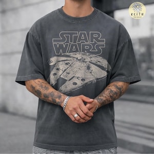 Pode incluir: Uma t-shirt de manga curta em cinzento-carvão com um gráfico bege da nave espacial Millennium Falcon e o texto STAR WARS numa fonte de contorno arrojada. A peça apresenta um aspeto desbotado e um decote redondo clássico.