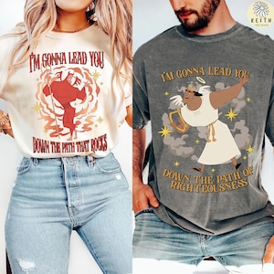 Puede incluir: Dos camisetas gráficas con ilustraciones de dibujos animados y texto. Una es de color crema con una figura de diablo rojo y las palabras "I'M GONNA LEAD YOU DOWN THE PATH THAT ROCKS." La otra es gris con un ángel y las palabras "I'M GONNA LEAD YOU DOWN THE PATH OF RIGHTEOUSNESS."