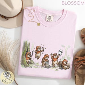 May include: A light pink Comfort Colors t-shirt featuring a whimsical illustration of Ewoks in a forest setting. The design includes a Stormtrooper helmet and the text "BLOSSOM" at the top. A gold necklace and sandals are also visible.