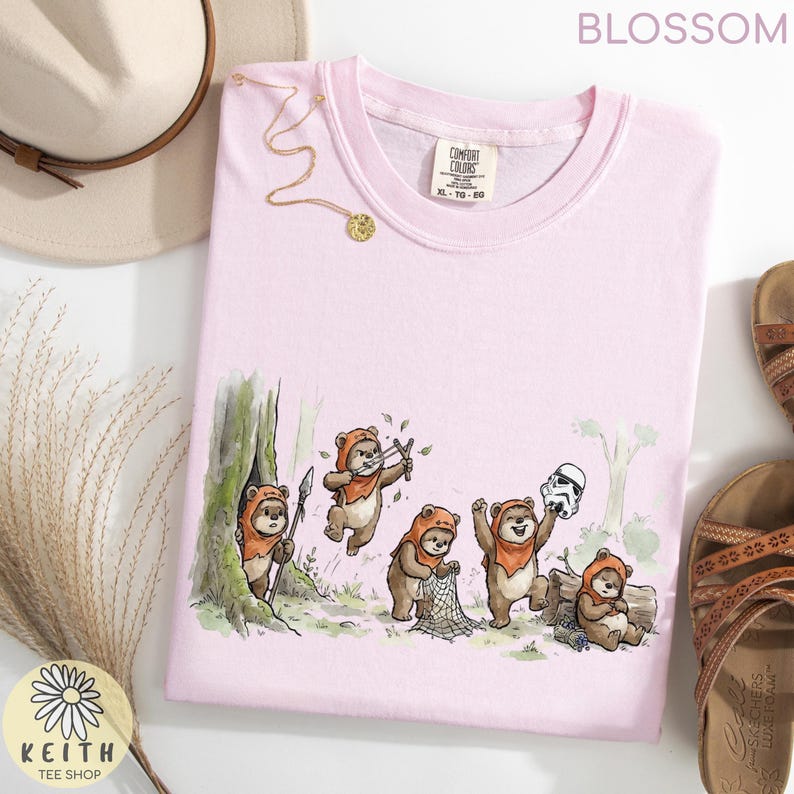 May include: A light pink Comfort Colors t-shirt featuring a whimsical illustration of Ewoks in a forest setting. The design includes a Stormtrooper helmet and the text "BLOSSOM" at the top. A gold necklace and sandals are also visible.