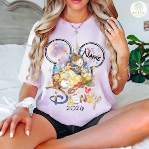 May include: A light purple t-shirt featuring a Disney design with the text "Disney 2026". The design includes the characters Belle, Beast, and other characters from Beauty and the Beast. The shirt has a Mickey Mouse ear design with the word "Name" above the characters.