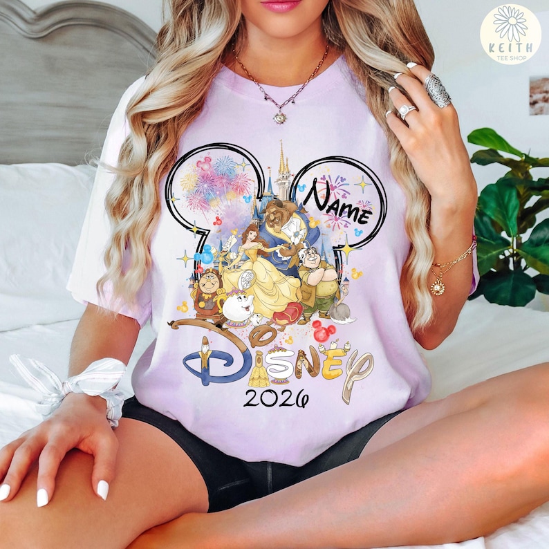 May include: A light purple t-shirt featuring a Disney design with the text "Disney 2026". The design includes the characters Belle, Beast, and other characters from Beauty and the Beast. The shirt has a Mickey Mouse ear design with the word "Name" above the characters.